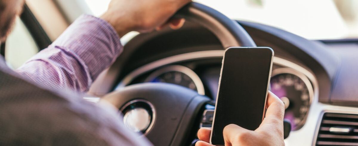 Ilustrační stock: řidič drží v ruce mobilní telefon za volantem osobního automobilu, pohled zprava nebo zleva na interiér vozu během jízdy, civilně, bez loga výrobce, fotoreal
