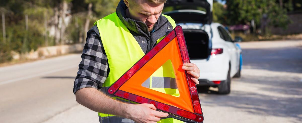 Ilustrační stock: řidič v reflexní vestě postavil výstražný trojúhelník na okraj silnice, za ním stojí auto s rozsvícenými výstražnými světly, civilně